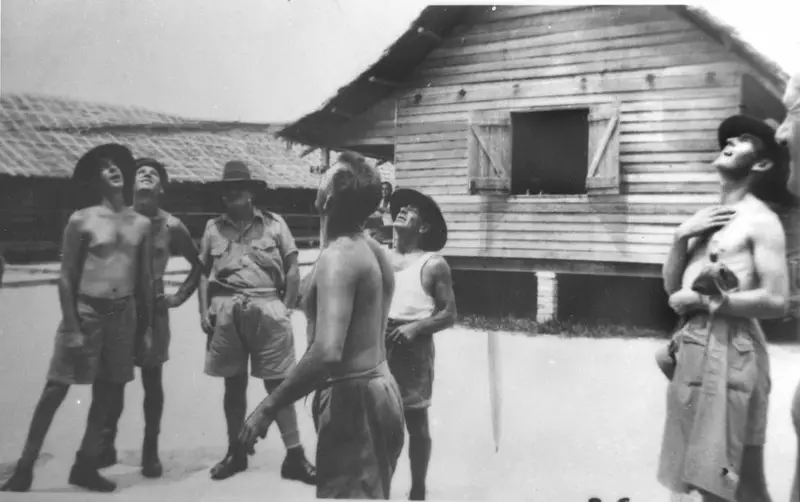 A ring of Australian soldiers watching two pennies in the air outside wooden army huts in 1941